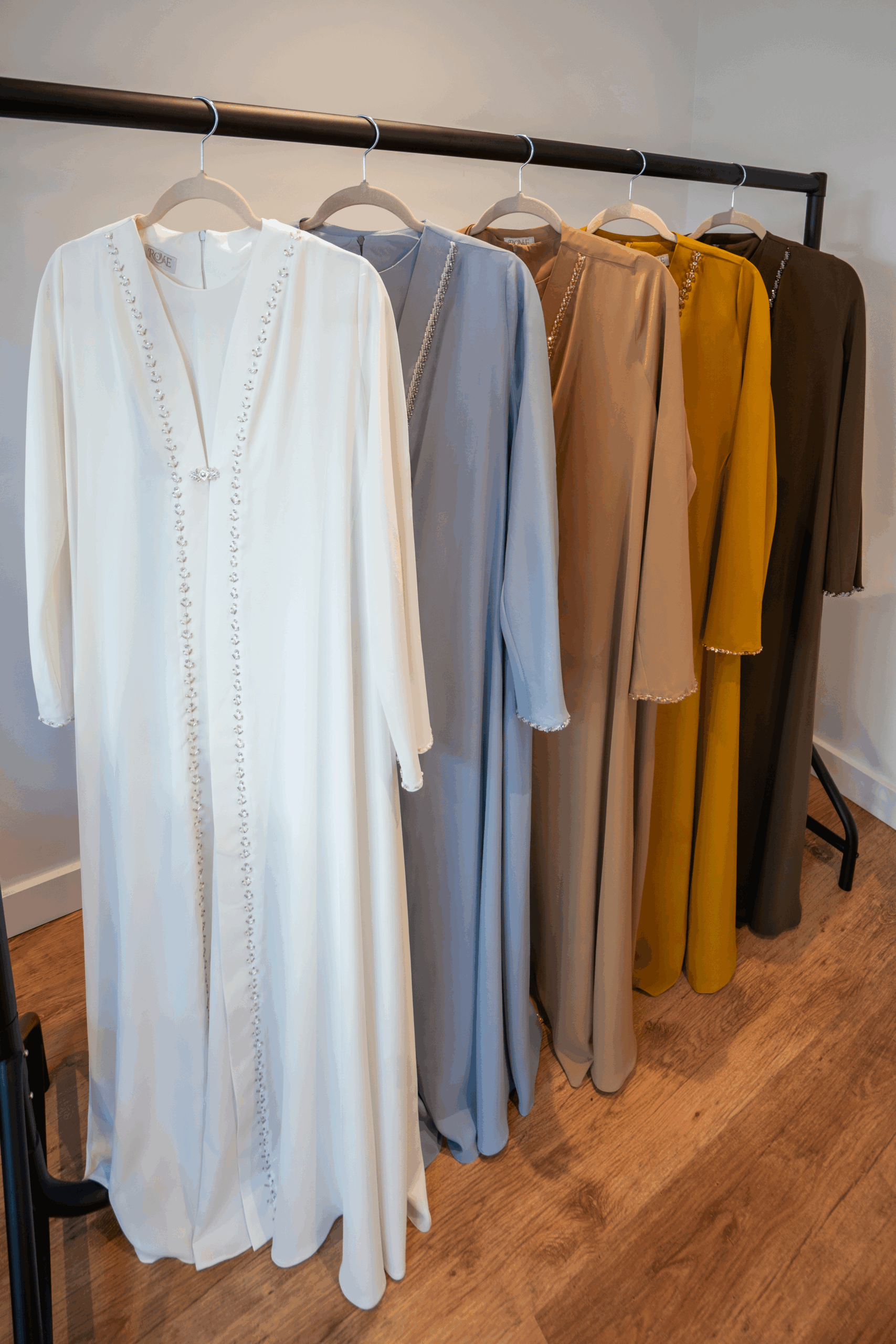 All five colours of the Adoria Abaya collection displayed on a clothing rail – white, cream camel, baby blue, mustard and dark brown.