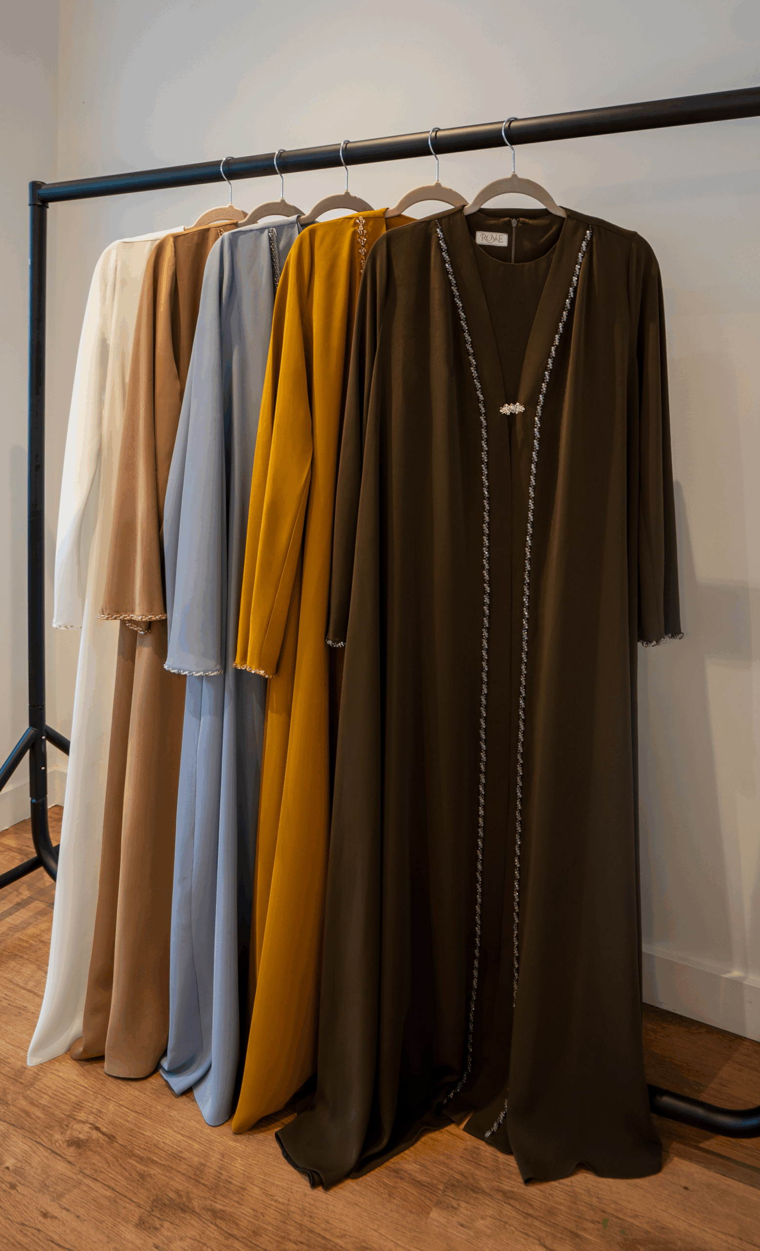 Five Royaé ADORIA hand-embellished abayas in white, cream camel, baby blue, mustard and dark brown displayed on a clothing rail.